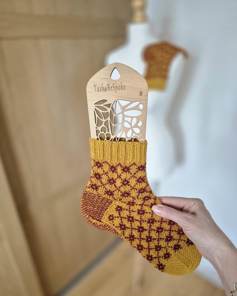 May include: Mustard yellow and burgundy patterned sock held in front of a wooden display with the text "TashaBeSpoke". The sock has a geometric design and is displayed on a wooden form. A matching knitted garment is in the background.