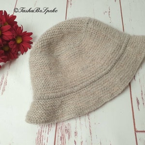 May include: A light beige knitted bucket hat with a slightly curved brim. The hat is made of a textured yarn and sits on a white and light brown wooden surface. Red flowers are in the upper left corner. The text "TashaBeSpoke" is in the upper left corner.