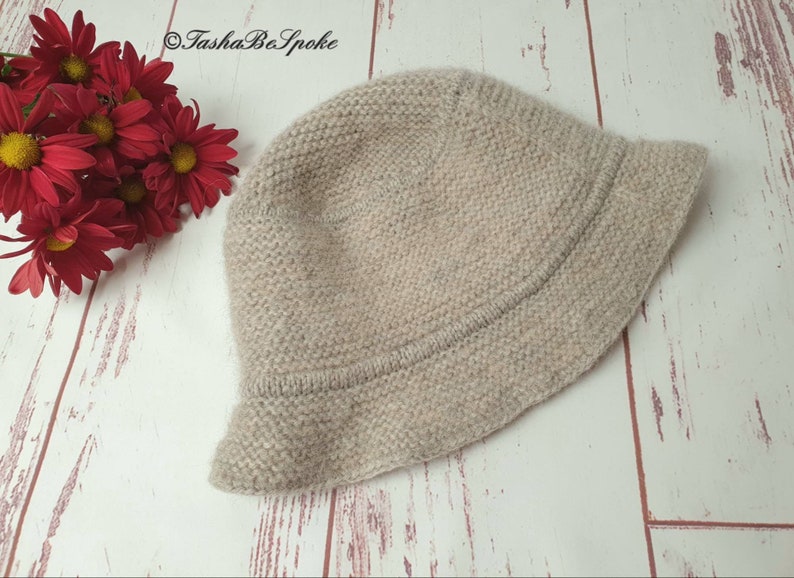 May include: A light beige knitted bucket hat with a slightly curved brim. The hat is made of a textured yarn and sits on a white and light brown wooden surface. Red flowers are in the upper left corner. The text "TashaBeSpoke" is in the upper left corner.