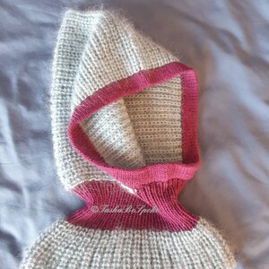 Pu&ograve; includere: Un cappuccio lavorato a mano a maglia grigio e magenta. Il cappuccio &egrave; realizzato con un filato morbido e strutturato con un design a coste. Ha un bordo magenta intorno al viso e al collo. La parte inferiore del cappuccio si allarga leggermente. Il testo "TashaBeSpoke" &egrave; visibile.