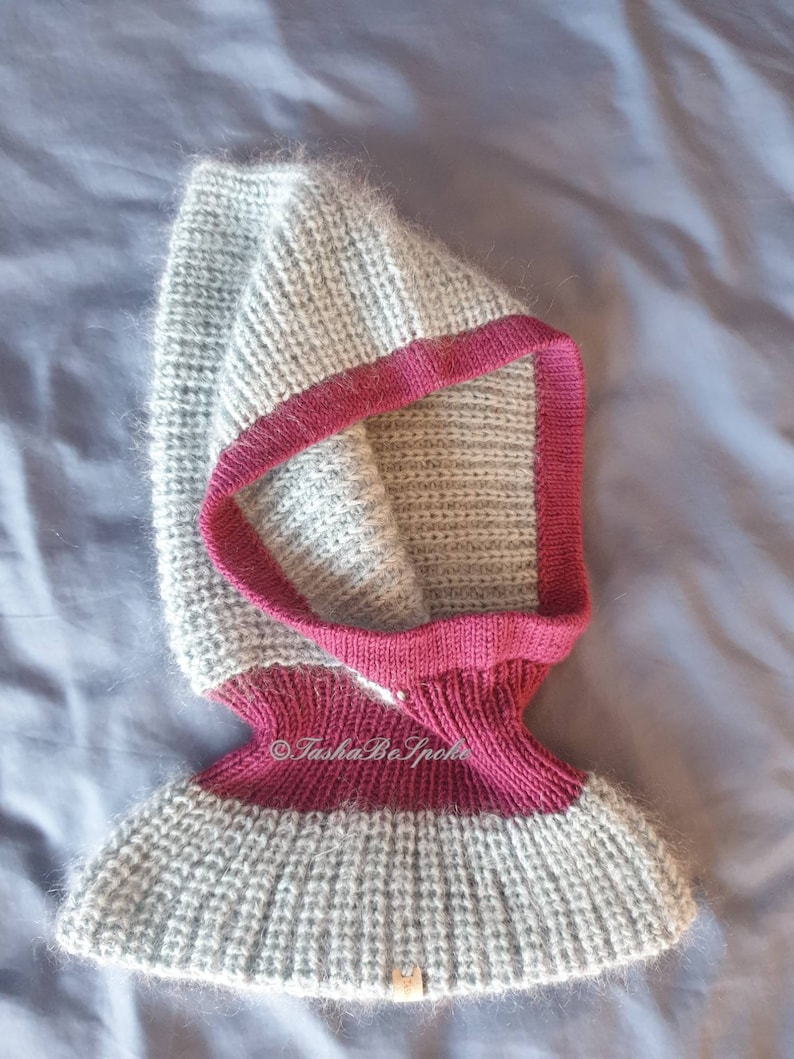 Pu&ograve; includere: Un cappuccio lavorato a mano a maglia grigio e magenta. Il cappuccio &egrave; realizzato con un filato morbido e strutturato con un design a coste. Ha un bordo magenta intorno al viso e al collo. La parte inferiore del cappuccio si allarga leggermente. Il testo "TashaBeSpoke" &egrave; visibile.