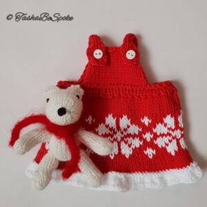 May include: A white knitted teddy bear wearing a red scarf, standing next to a red knitted dress with white snowflake pattern and white trim.