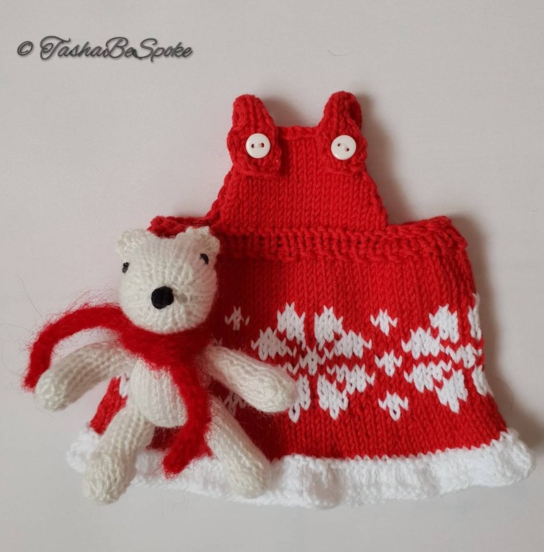 May include: A white knitted teddy bear wearing a red scarf, standing next to a red knitted dress with white snowflake pattern and white trim.