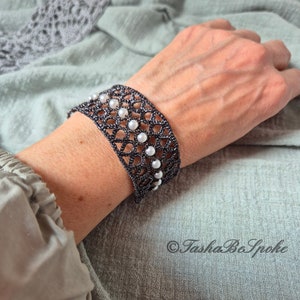 Puede incluir: Un brazalete de crochet negro con detalles de perlas blancas. El brazalete est&aacute; en la mu&ntilde;eca de una persona.