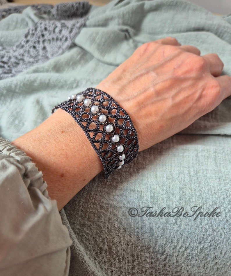 Puede incluir: Un brazalete de crochet negro con detalles de perlas blancas. El brazalete est&aacute; en la mu&ntilde;eca de una persona.