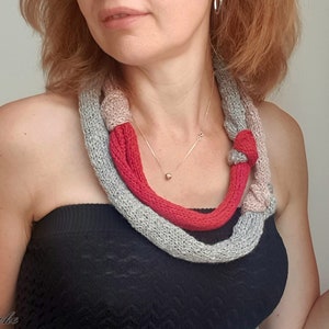 May include: A knitted necklace in shades of gray, red, and beige. The necklace is knotted in several places and is worn around the neck. A gold necklace is also visible.