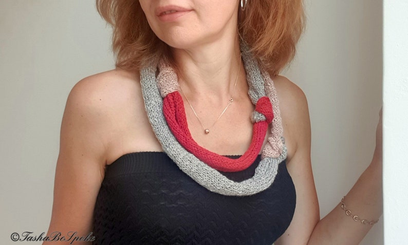 May include: A knitted necklace in shades of gray, red, and beige. The necklace is knotted in several places and is worn around the neck. A gold necklace is also visible.