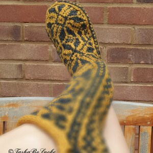 May include: A pair of long, knitted socks in a mustard yellow and black color scheme with a geometric pattern. The socks are worn on a person's legs and the foot of one sock is visible.