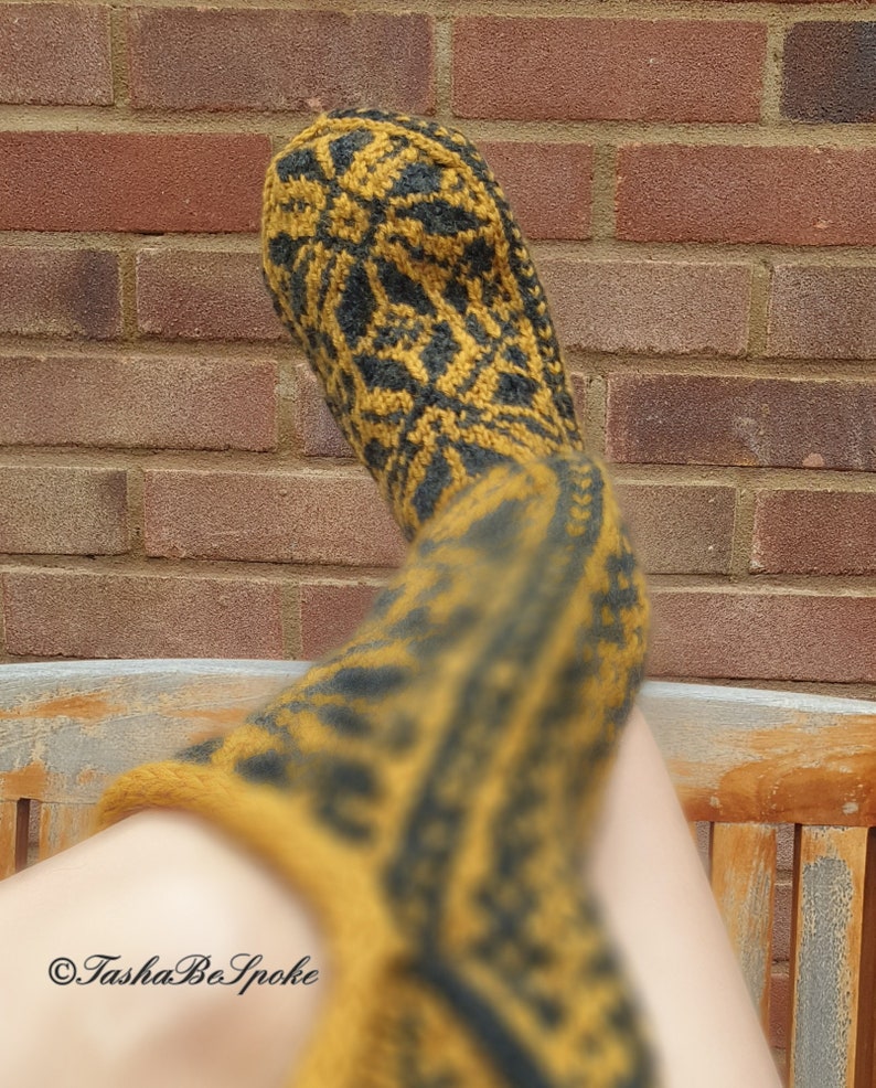 May include: A pair of long, knitted socks in a mustard yellow and black color scheme with a geometric pattern. The socks are worn on a person's legs and the foot of one sock is visible.