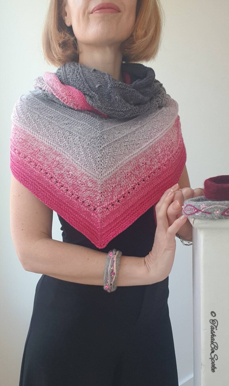 May include: A grey, pink and white triangular shawl with a lacy pattern. The shawl is being worn by a person with short blonde hair. The person is wearing a black top and a bracelet.