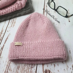 K&ouml;nnte beinhalten: Eine rosafarbene Strickm&uuml;tze mit einem braunen Lederlabel, auf dem "TashaBeSpoke" steht.