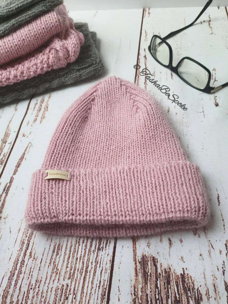 K&ouml;nnte beinhalten: Eine rosafarbene Strickm&uuml;tze mit einem braunen Lederlabel, auf dem "TashaBeSpoke" steht.