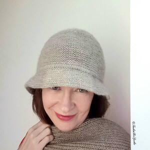 May include: A light gray knitted cloche hat with a small brim. The hat is made of a soft, textured yarn. The hat is shown on a person wearing a matching scarf.