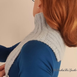 May include: A light blue knitted cowl neck warmer with a ribbed texture. The cowl is being worn by a person with long red hair.