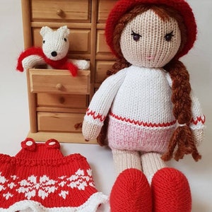 May include: A knitted doll with brown hair wearing a red hat, white sweater, pink skirt, and red shoes. The doll is holding a small white knitted animal wearing a red scarf. The doll is sitting in front of a wooden dresser with multiple drawers.