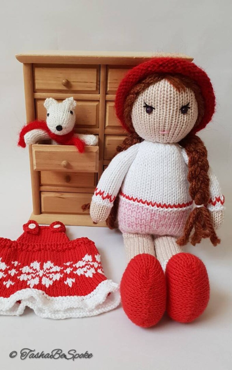 May include: A knitted doll with brown hair wearing a red hat, white sweater, pink skirt, and red shoes. The doll is holding a small white knitted animal wearing a red scarf. The doll is sitting in front of a wooden dresser with multiple drawers.