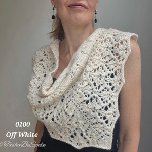 May include: A white crocheted shawl with a delicate lace pattern. The shawl is draped over a person's shoulders and is made with a soft, lightweight yarn. The shawl is labelled "0100 Off White" and "Tasha Be Spoke".