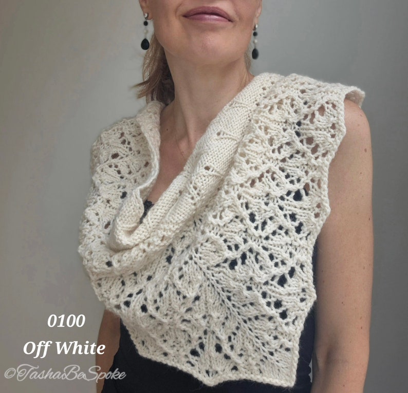 May include: A white crocheted shawl with a delicate lace pattern. The shawl is draped over a person's shoulders and is made with a soft, lightweight yarn. The shawl is labelled "0100 Off White" and "Tasha Be Spoke".