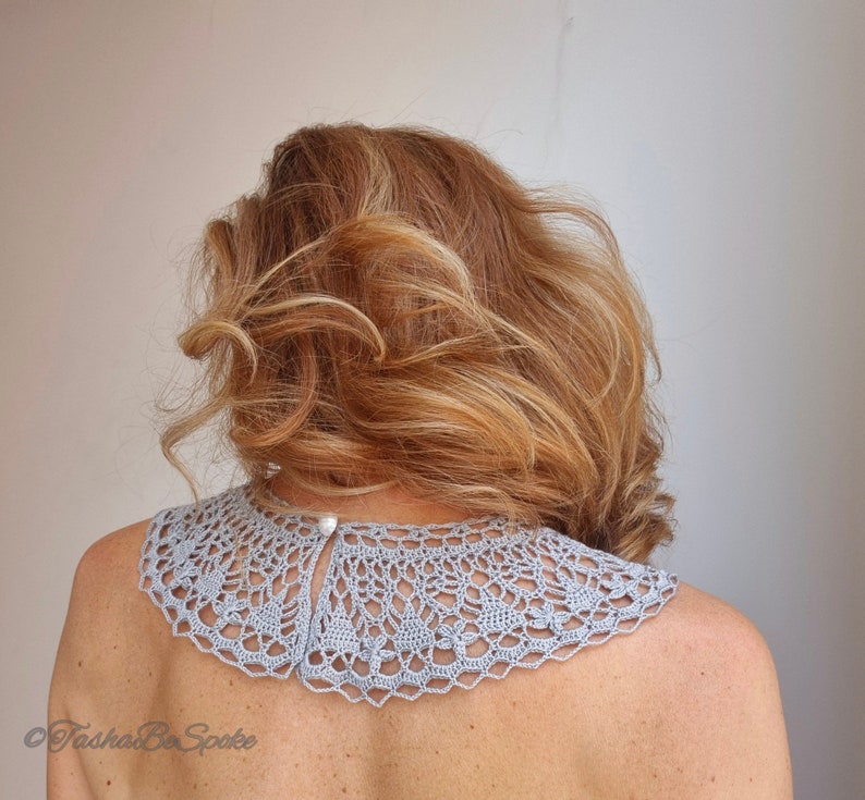 May include: A light gray crocheted collar with a delicate pattern. The collar is worn by a person with long, wavy, light brown hair.