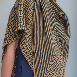 Pode incluir: Um chal triangular com um padr&atilde;o geom&eacute;trico em tons de amarelo mostarda e azul turquesa. O chal &eacute; tricotado com um fio fino e tem uma franja delicada.