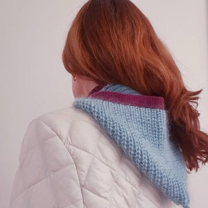Pu&ograve; includere: Un collo a maglia blu chiaro e viola. La sciarpa &egrave; indossata sopra una giacca trapuntata bianca. La modella ha lunghi capelli rossi. La sciarpa &egrave; a forma di triangolo e sembra fatta a mano.