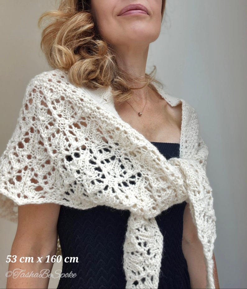 May include: A white crocheted shawl with a delicate lace pattern. The shawl is 53 cm x 160 cm and is shown draped around a person's shoulders.