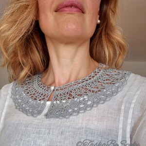 May include: A gray crocheted collar with a pearl button closure. The collar is worn over a white linen shirt.