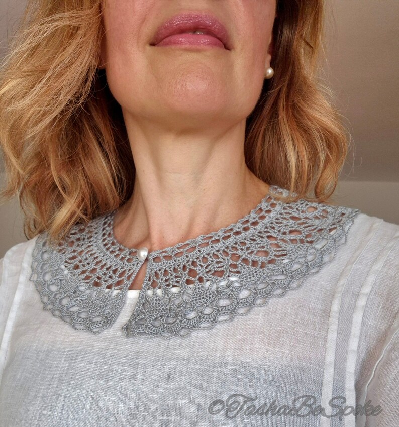 May include: A gray crocheted collar with a pearl button closure. The collar is worn over a white linen shirt.