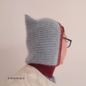 Pu&ograve; includere: Un cappuccio lavorato a maglia a mano, blu chiaro, con una parte superiore a punta e un bordo bordeaux. Il cappuccio &egrave; progettato per coprire la testa e il collo, offrendo calore e protezione. Il cappuccio &egrave; indossato sopra una giacca bianca.