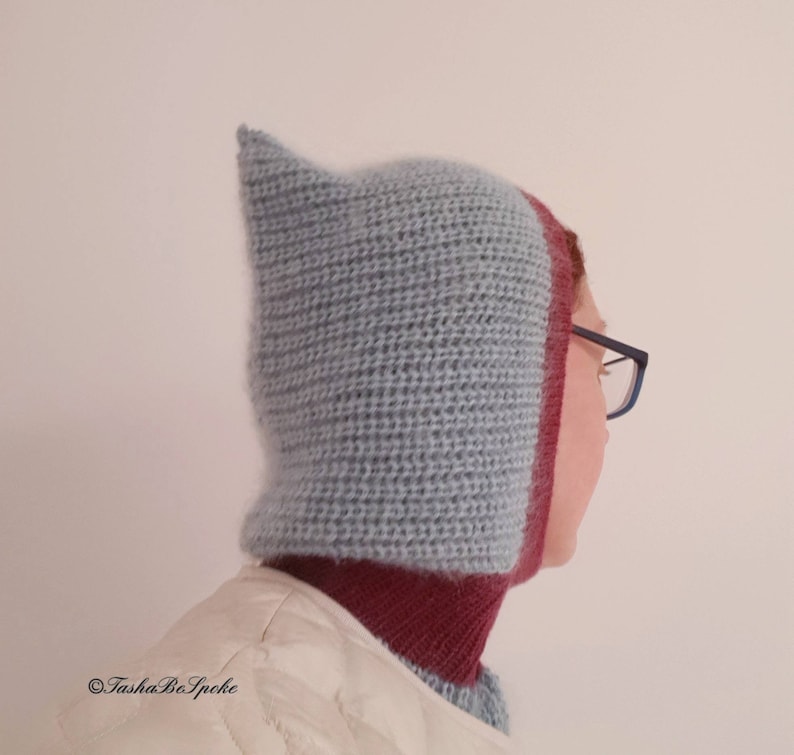Pu&ograve; includere: Un cappuccio lavorato a maglia a mano, blu chiaro, con una parte superiore a punta e un bordo bordeaux. Il cappuccio &egrave; progettato per coprire la testa e il collo, offrendo calore e protezione. Il cappuccio &egrave; indossato sopra una giacca bianca.