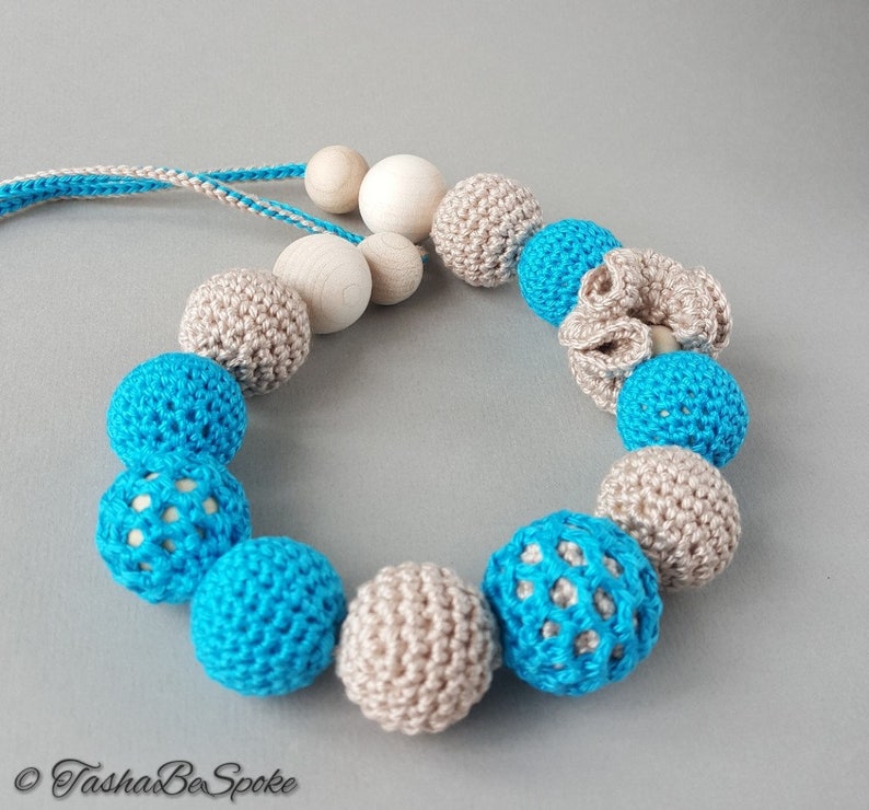 Op de afbeelding: Een handgemaakte ketting met een mix van gehaakte en houten kralen. De ketting heeft een kleurenschema van lichtblauw, beige en natuurlijk hout. De gehaakte kralen zijn rond, sommige hebben een open, kanten ontwerp. De ketting is geregen op een blauw-wit koord.