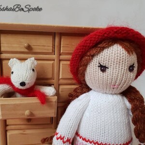 May include: A knitted doll with brown hair wearing a red hat and white sweater. The doll is holding a white knitted animal wearing a red scarf.