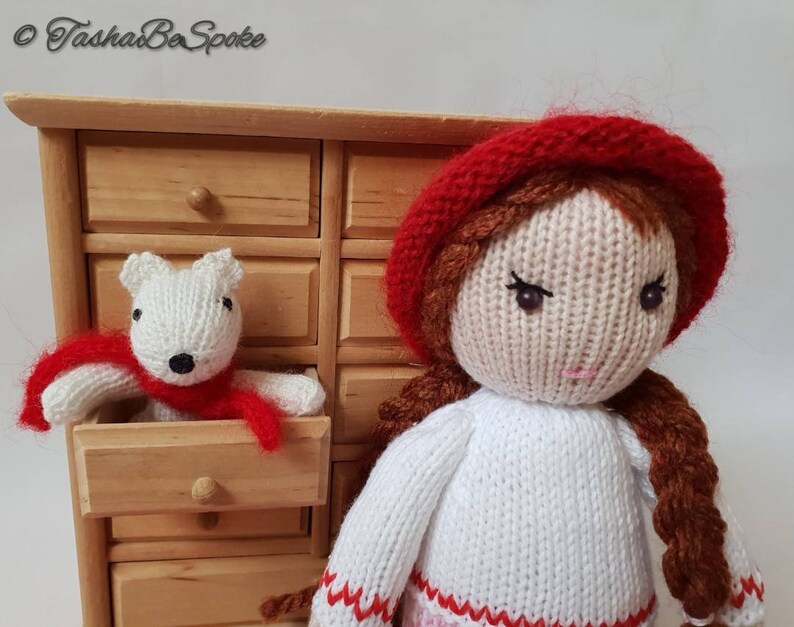 May include: A knitted doll with brown hair wearing a red hat and white sweater. The doll is holding a white knitted animal wearing a red scarf.