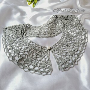 May include: A gray crocheted collar with a pearl button in the center. The collar is laid out on a white satin fabric.