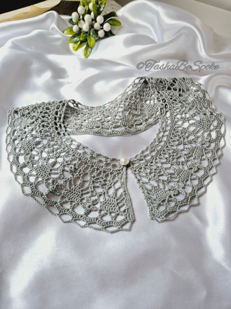 May include: A gray crocheted collar with a pearl button in the center. The collar is laid out on a white satin fabric.