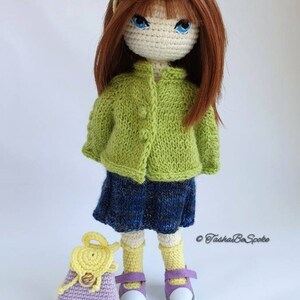 Pu&ograve; includere: Una bambola all'uncinetto con capelli castani, un cardigan verde, una gonna blu, calzini gialli e scarpe viola. La bambola tiene uno zaino all'uncinetto viola e giallo.
