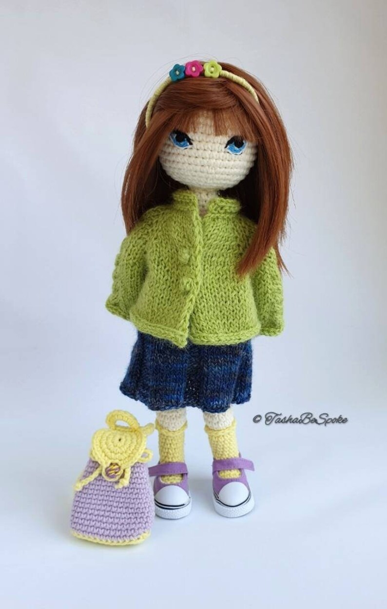 Pu&ograve; includere: Una bambola all'uncinetto con capelli castani, un cardigan verde, una gonna blu, calzini gialli e scarpe viola. La bambola tiene uno zaino all'uncinetto viola e giallo.