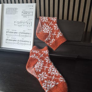 May include: A pair of orange and white snowflake patterned socks. The socks are knitted with a ribbed cuff, heel, and toe. The socks are resting on a dark surface near a music book.