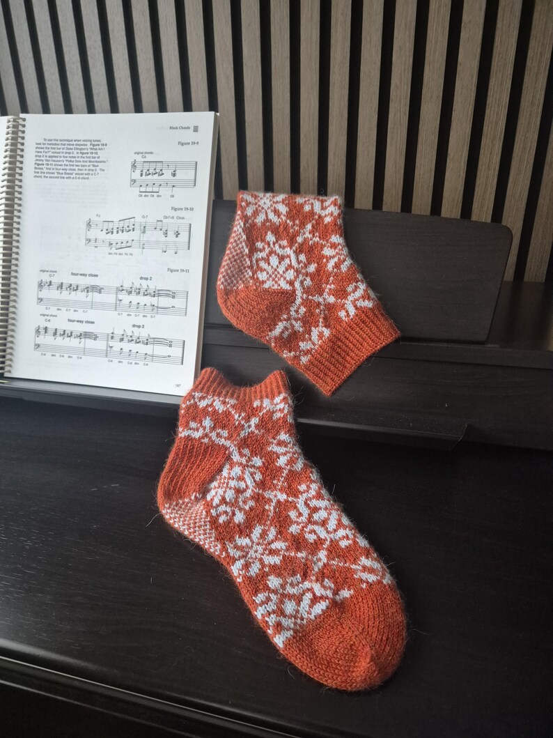 May include: A pair of orange and white snowflake patterned socks. The socks are knitted with a ribbed cuff, heel, and toe. The socks are resting on a dark surface near a music book.