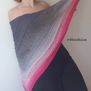 May include: A grey, pink and white ombre knitted shawl with a lacy pattern. The shawl is draped over a person's shoulders and is being held up by their right hand. The text "&copy; TashaBeSpoke" is in the bottom right corner of the image.