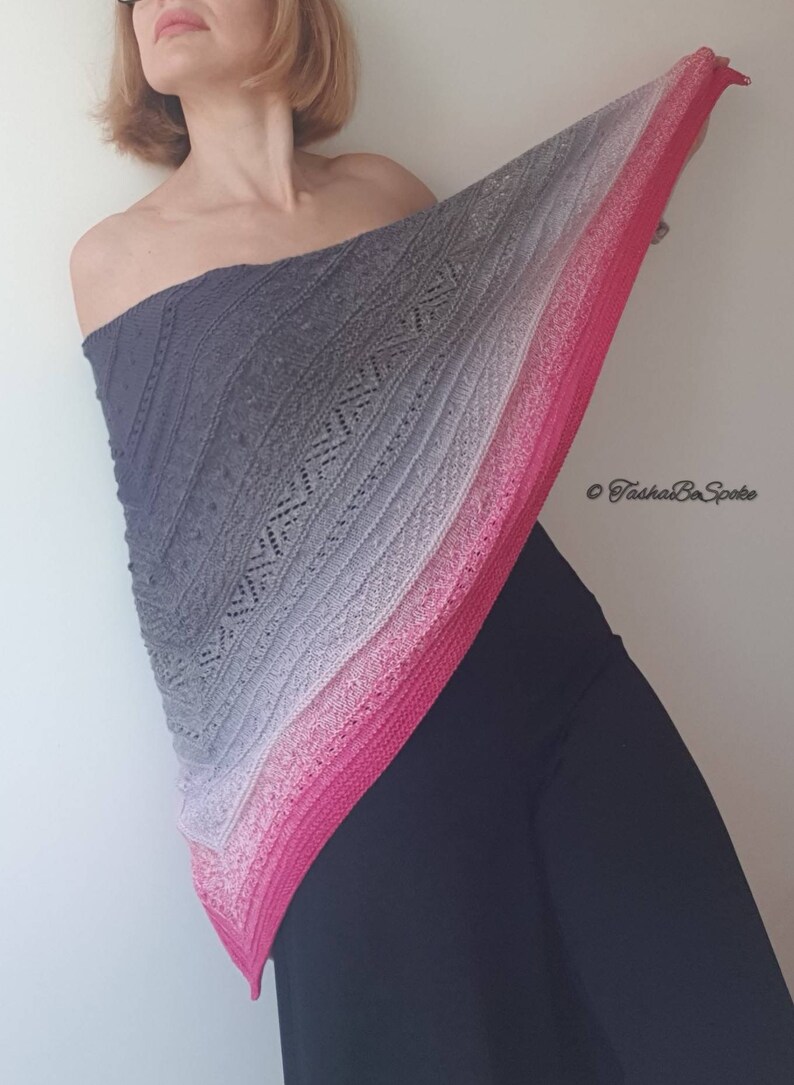 May include: A grey, pink and white ombre knitted shawl with a lacy pattern. The shawl is draped over a person's shoulders and is being held up by their right hand. The text "&copy; TashaBeSpoke" is in the bottom right corner of the image.