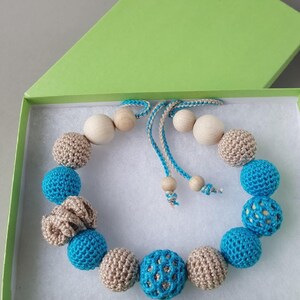 Op de afbeelding: Een handgemaakte ketting met afwisselend blauwe en bruine gehaakte kralen, afgewisseld met houten kralen. De ketting wordt gepresenteerd in een groene doos met een witte binnenkant, wat suggereert dat het een cadeauartikel is. Het ontwerp is eenvoudig en elegant.