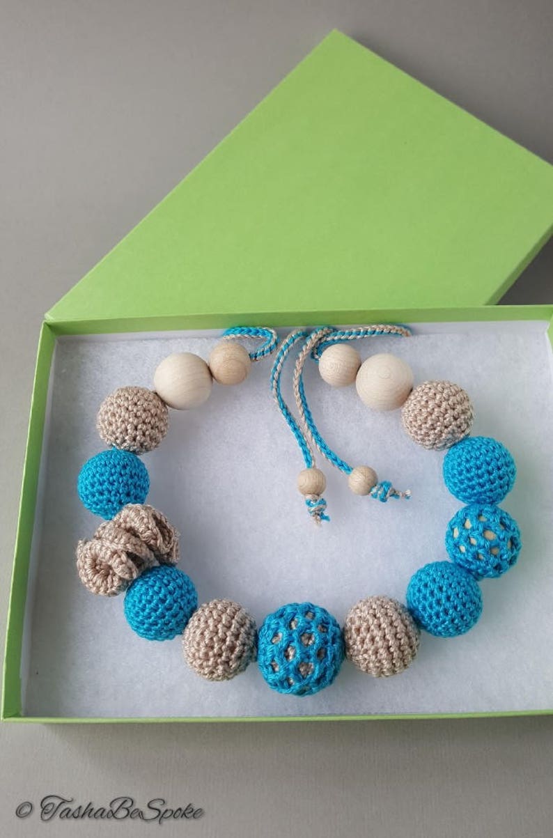 Op de afbeelding: Een handgemaakte ketting met afwisselend blauwe en bruine gehaakte kralen, afgewisseld met houten kralen. De ketting wordt gepresenteerd in een groene doos met een witte binnenkant, wat suggereert dat het een cadeauartikel is. Het ontwerp is eenvoudig en elegant.