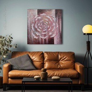 May include: A textured painting of a rose in white and pink hues, set against a striped brown and pink background. The artwork is displayed above a brown leather sofa. The room features a black coffee table.