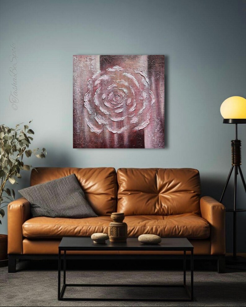 May include: A textured painting of a rose in white and pink hues, set against a striped brown and pink background. The artwork is displayed above a brown leather sofa. The room features a black coffee table.