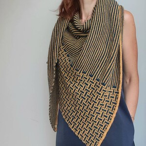 Pode incluir: Um chal triangular com um padr&atilde;o geom&eacute;trico em tons de amarelo mostarda e azul marinho. O chal &eacute; tricotado com um fio fino e tem uma franja delicada.