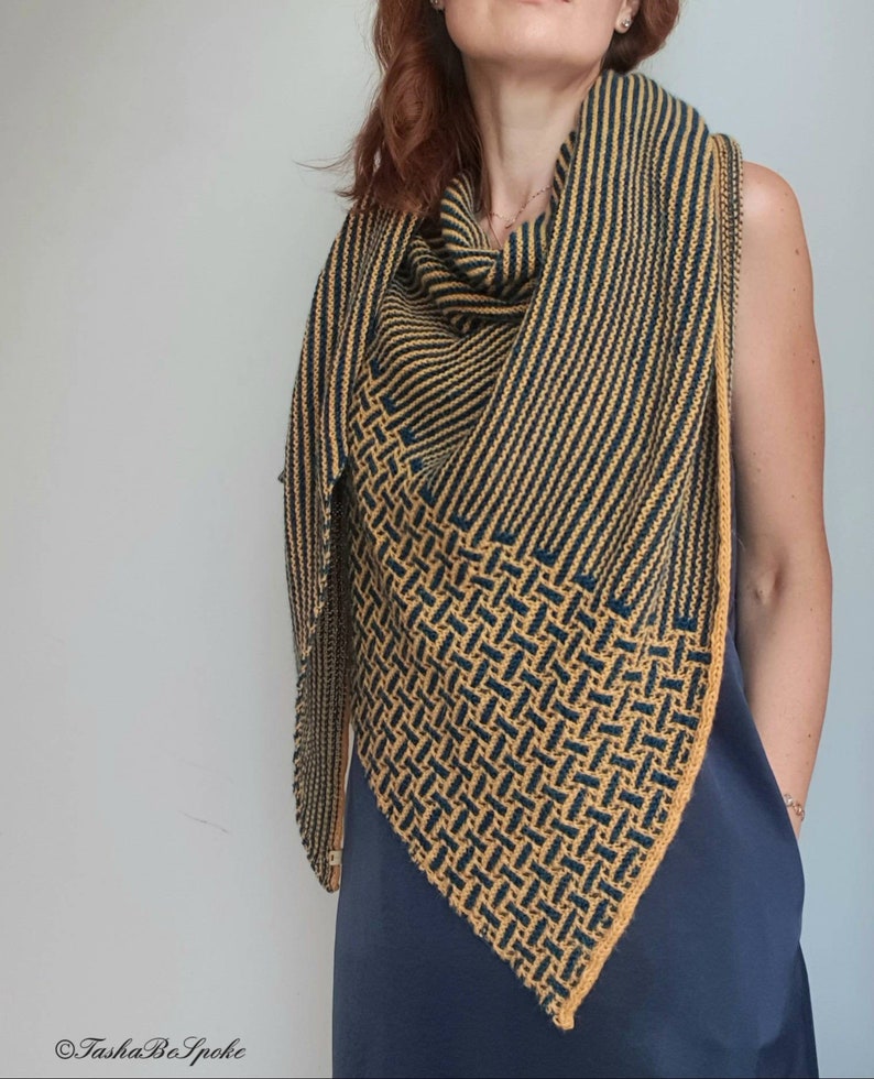 Pode incluir: Um chal triangular com um padr&atilde;o geom&eacute;trico em tons de amarelo mostarda e azul marinho. O chal &eacute; tricotado com um fio fino e tem uma franja delicada.