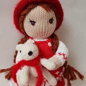 May include: A knitted doll wearing a red dress and a red hat, holding a white knitted teddy bear wearing a red scarf.