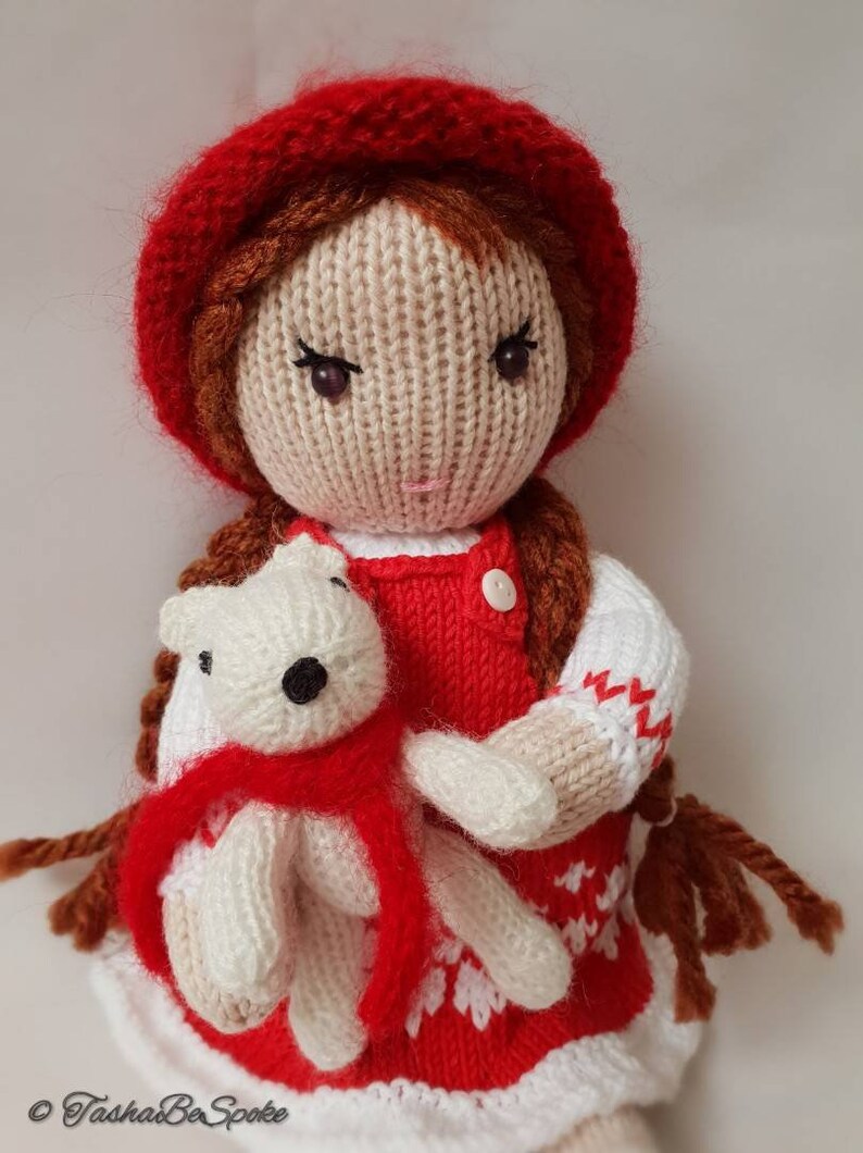 May include: A knitted doll wearing a red dress and a red hat, holding a white knitted teddy bear wearing a red scarf.
