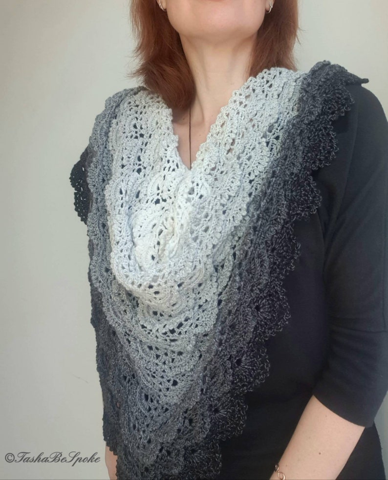 May include: A grey and black crocheted shawl with a scalloped edge. The shawl is made with a gradient yarn that transitions from light grey to dark grey.